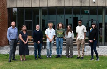 Members of the Pitch for the Planet judging panel and winning entrants stand in a line with the university campus as a backdrop