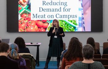 The Sustainability Manager, Sam Waugh, talks in front of an audience. The screen behind her shows a slide that reads 