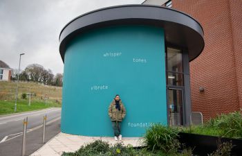 Helen Cammock stands in front of the artwork on an external wall at top of University of Sussex Student Centre. The text is painted in white and set against a bright teal background, and reads: whisper  tones  vibrate  foundations