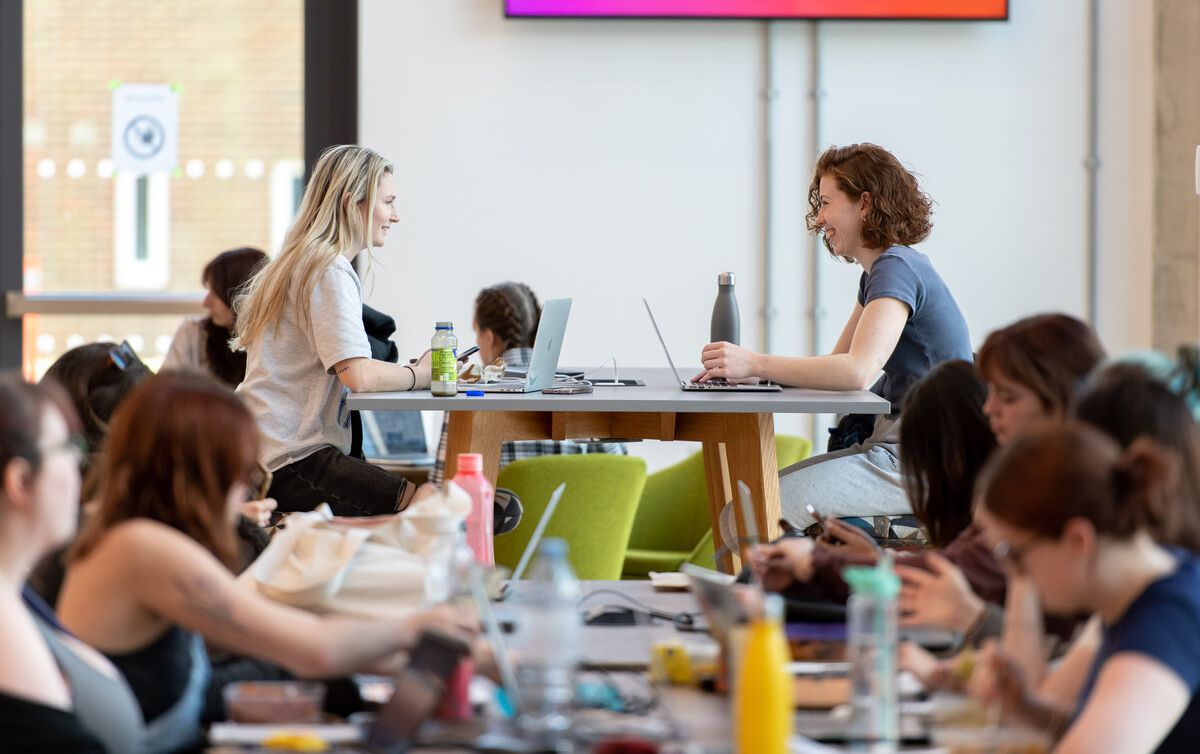 Two happy students sat at a table with their laptops in the student centre